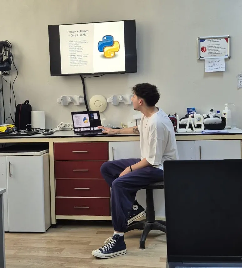 Presenting the project in the laboratory, seated at the bench with a laptop, the presentation slide mirrored on the wall-mounted display above.