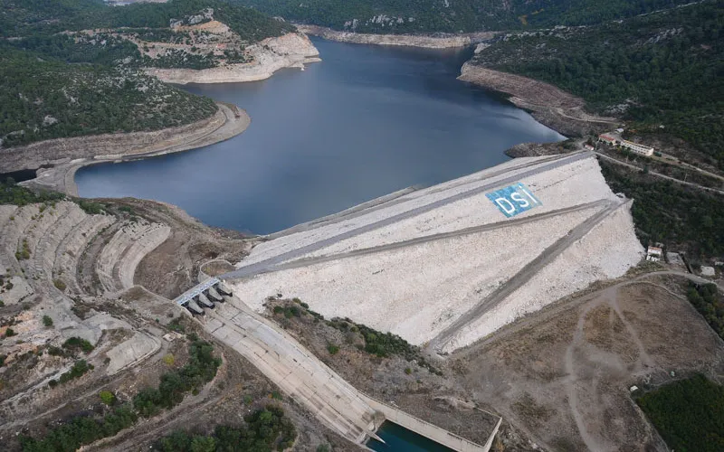 Aerial view of Tahtalı Dam, its concrete face marked DSİ, holding back a reservoir in wooded hills. This is one of the reservoirs the project models.